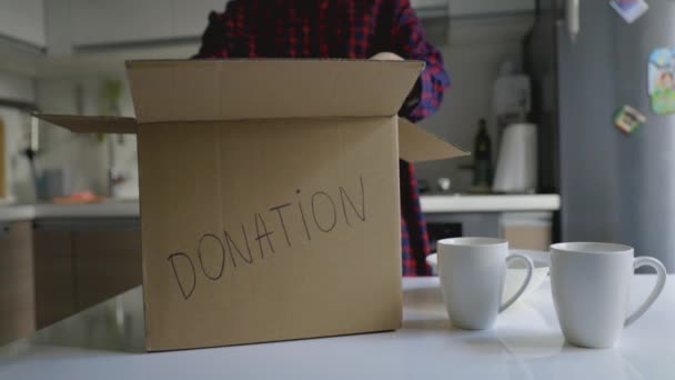 darovat předměty pro domácnost - žena dává stolní nádobí do kartonové krabičky k daru na kuchyňský stůl