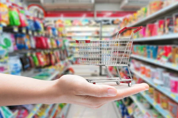 Mão segurando carrinho de compras com fundo de supermercado — Fotografia de Stock