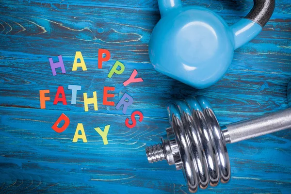 Happy Father\'s Day inscription with kettlebell and dumbbell on blue wooden table
