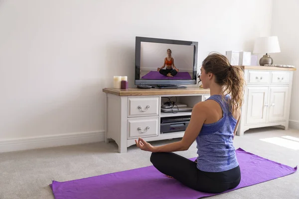 Genç, sağlıklı bir kadın TV karşısında yoga yapıyor.