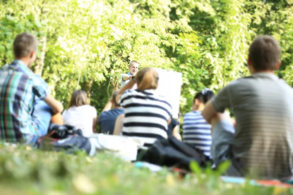Um grupo de jovens estudantes no parque. Vista de um homem gesticulando com as mãos, em pé contra um grupo desfocado de pessoas sentadas na frente dele na grama . — Fotografia de Stock