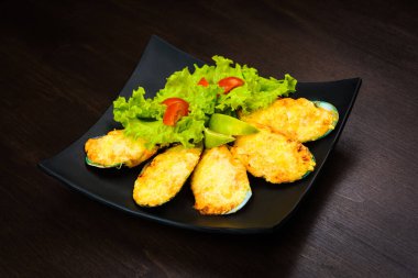 Peynirli fırında istiridye, marul ve domates, ahşap kahverengi masada, siyah bir tabakta.