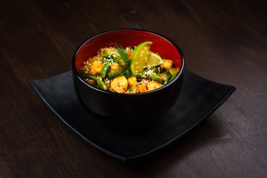 Kahverengi ahşap arka planda siyah yuvarlak kâsede karidesli sebze tavası.