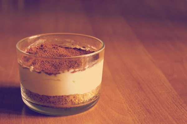 Bolo de queijo de leite em vidro, decorado com migalhas de café, em uma mesa de madeira em um fundo desfocado no início da manhã de verão — Fotografia de Stock