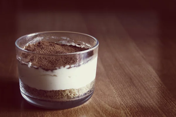 Milk cheesecake in glass, decorated with coffee crumbs, on a wooden table on a blurred background in the early summer morning — Stock Photo, Image