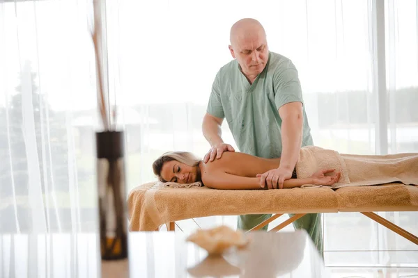 Blond kvinna avkopplande få ryggmassage från manlig sjukgymnast i skönhetssalong ligger på massagebord. Ung flicka med tatueringar avkopplande i spa center koncept - Medium — Stockfoto