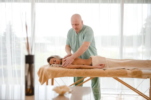 Blonde woman relaxing receiving back massage from male physiotherapist in beauty salon lying on massage table. Young girl with tattoos relaxing in spa center concept - Medium