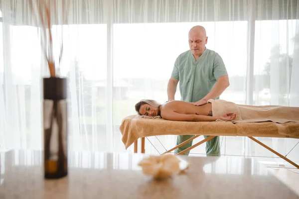 Blond kvinna avkopplande få ryggmassage från manlig sjukgymnast i skönhetssalong ligger på massagebord. Ung flicka med tatueringar avkopplande i spa center koncept - Medium — Stockfoto