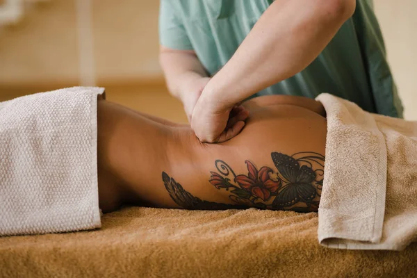 Mujer rubia relajante recibiendo masaje de espalda de fisioterapeuta masculino en el salón de belleza acostado en la mesa de masaje. Chica joven con tatuajes relajante en concepto de centro de spa - Medio — Foto de Stock