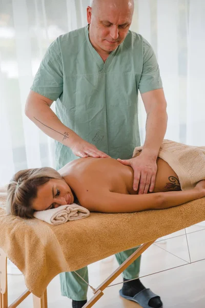 Blonde woman relaxing receiving back massage from male physiotherapist in beauty salon lying on massage table. Young girl with tattoos relaxing in spa center concept - Close-up