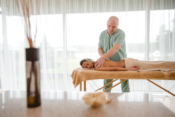 Блондинка розслабляється, отримуючи масаж спини від чоловіка-фізіотерапевта в салоні краси, що лежить на масажному столі. Молода дівчина з татуюваннями розслабляється в концепції спа-центру - Medium — стокове фото