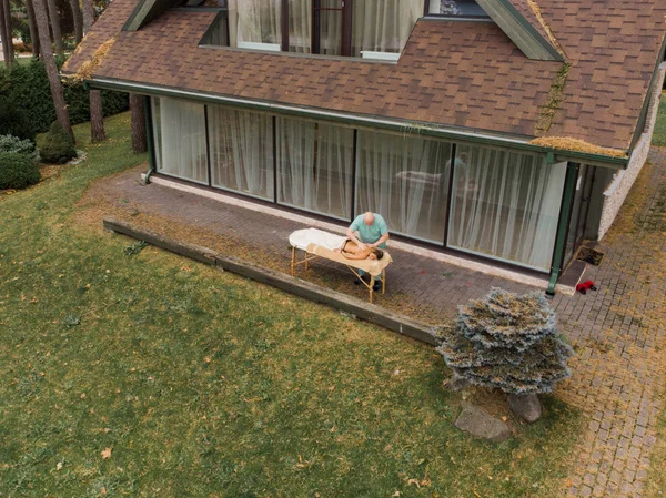 AERIAL: Blonde woman relaxing receiving back massage from male physiotherapist outdoor in wood country house lying on massage table. Young girl with tattoos relaxing destination