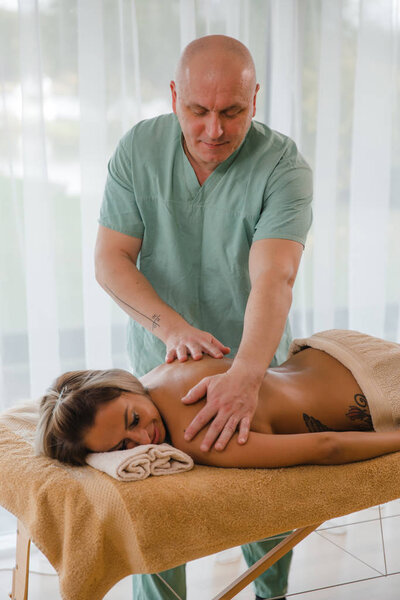 Blonde woman relaxing receiving back massage from male physiotherapist in beauty salon lying on massage table. Young girl with tattoos relaxing in spa center concept - Close-up