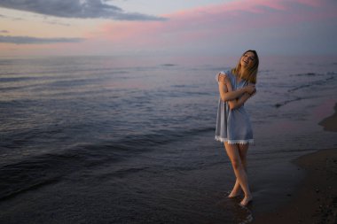 Gün batımında Baltık Denizi sahilinde açık mavi elbiseli güzel bir sarışının portresi canlı renklerle
