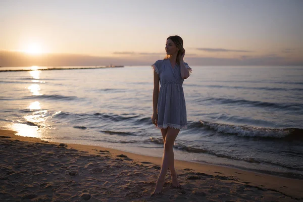 De pie a toda altura: Retrato de una hermosa mujer rubia en un vestido azul claro en la playa del Mar Báltico durante la puesta de sol con colores vivos —  Fotos de Stock