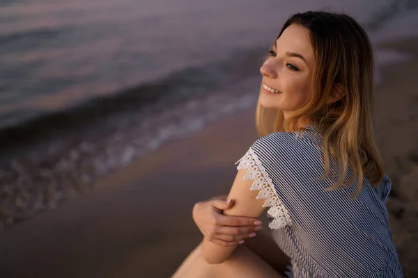 Sentado: Retrato de uma bela mulher loira em um vestido azul claro na praia do Mar Báltico durante o pôr do sol com cores vivas — Fotografia de Stock