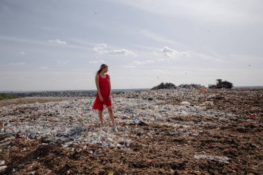Doğayı kirletme eylemcisi, dışarıdaki büyük bir çöplükte kırmızı elbiseli genç sarışın kadın dünyamızdaki tüm insan atıklarına ve plastiklerine bakıyor Doğu Avrupa 'da Letonya Riga' da uçan martılar