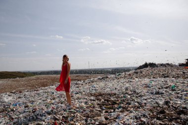 Doğayı kirletme eylemcisi, dışarıdaki büyük bir çöplükte kırmızı elbiseli genç sarışın kadın dünyamızdaki tüm insan atıklarına ve plastiklerine bakıyor Doğu Avrupa 'da Letonya Riga' da uçan martılar