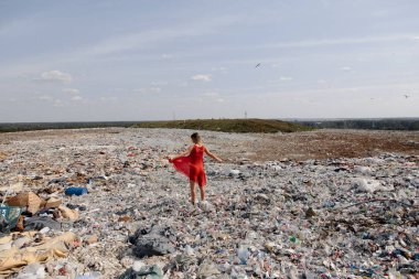 Doğayı kirletme eylemcisi, dışarıdaki büyük bir çöplükte kırmızı elbiseli genç sarışın kadın dünyamızdaki tüm insan atıklarına ve plastiklerine bakıyor Doğu Avrupa 'da Letonya Riga' da uçan martılar