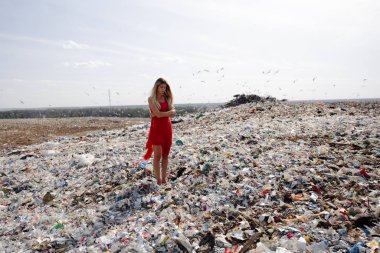 Doğayı kirletme eylemcisi, dışarıdaki büyük bir çöplükte kırmızı elbiseli genç sarışın kadın dünyamızdaki tüm insan atıklarına ve plastiklerine bakıyor Doğu Avrupa 'da Letonya Riga' da uçan martılar