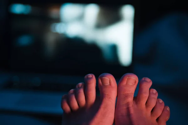 Primo piano di dita dell'uomo con unghie lunghe non tagliate che hanno urgente bisogno di pedicure - Due piedi seduti accanto a un computer portatile di notte con luci blu vivide al neon rosse e giallastre — Foto Stock