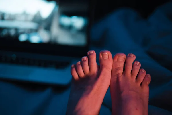 Detailní záběr na mužské prsty s dlouhými nehty, které naléhavě potřebují pedikúru - Dvě stopy sedí v noci vedle notebooku s neonově jasně modrými červenými a nažloutlými světly — Stock fotografie