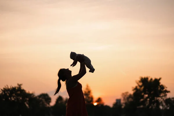 Sunset Siluet: Genç anne şehir parkı batan güneş ve canlı turuncu gökyüzü önünde duran onun bebek erkek çocuk tutan - Aile değerleri sıcak renk yaz sahnesi el
