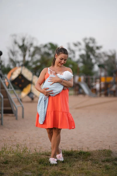 Allaitement : Jeune mère allaite son bébé garçon enfant dans le parc de la ville debout portant une robe rouge vif - Fils porte une casquette blanche - Valeurs familiales couleur chaude scène d'été portable — Photo