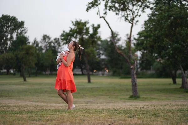 Jeune mère tenant et jouant avec son bébé garçon enfant dans le parc de la ville debout portant une robe rouge vif - Fils porte une casquette blanche - Valeurs familiales couleur chaude scène d'été portable — Photo