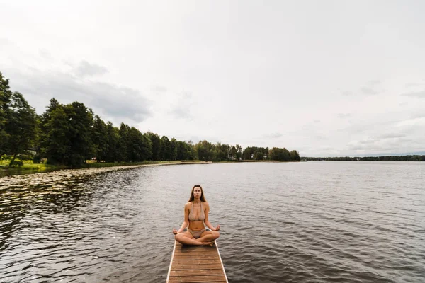 Fitte schlanke Frau praktiziert Yoga-Übungen im Mini-Badeanzug-Bikini am wolkenverhangenen See - Yoga-Meditation und Wellness-Lifestylekonzept - Volles Sitzen — Stockfoto