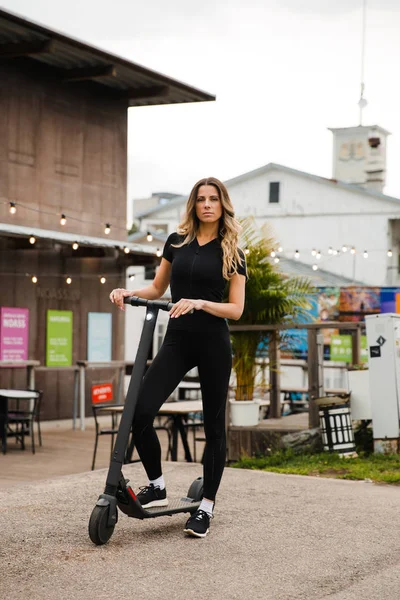 Fit Woman stands with electric scooter, rides away - Shot of Modern transportation gadget and popular futuristic device among young people - Eastern European Latvia Riga — Stock Photo, Image