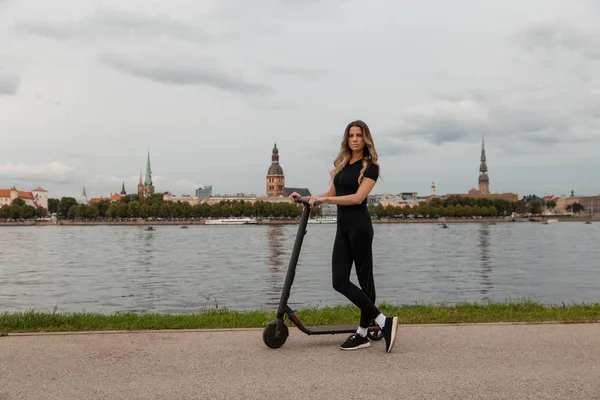 Passform kvinna av elektrisk skoter med en flod stad bakgrund - Skott av moderna transporter gadget och populära futuristiska enheten bland ungdomar - Östeuropa Lettland Riga — Stockfoto