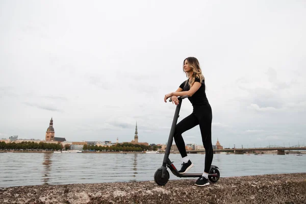 Passform kvinna av elektrisk skoter med en flod stad bakgrund - Skott av moderna transporter gadget och populära futuristiska enheten bland ungdomar - Östeuropa Lettland Riga — Stockfoto