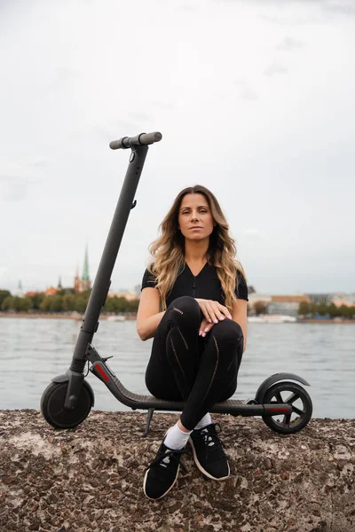 Nehir kenti geçmişine sahip elektrikli scooter kullanan fit bir kadın - Modern ulaşım araçları ve gençler arasında popüler fütürist cihazlar - Doğu Avrupa Letonya Riga — Stok fotoğraf