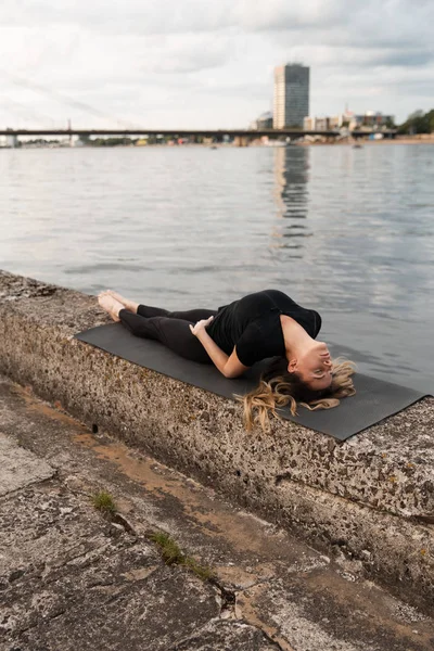 Yoga meditation and wellness lifestyle concept. Fit slim woman practicing yoga exercises near river Daugava and Vecriga old town background at sunset - Full sitting shot and meditating on rock beach