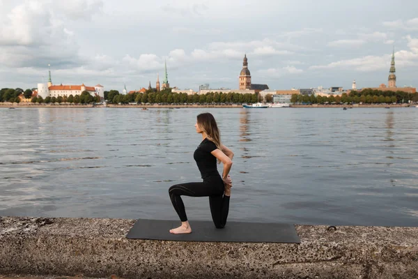 Yoga meditation and wellness lifestyle concept. Fit slim woman practicing yoga exercises near river Daugava and Vecriga old town background at sunset - Full sitting shot and meditating on rock beach