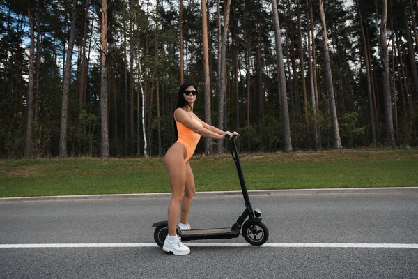 Otoparkta elektrikli scooter kullanan seksi bir kadın mayo ve güneş gözlüğü takıyor. Emisyonsuz çevre dostu ulaşım.