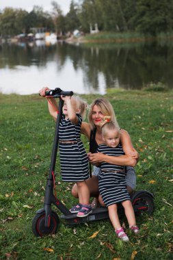 İkiz sarışın küçük kızlar parktaki elektrikli scooter 'da anneleriyle oturuyorlar.