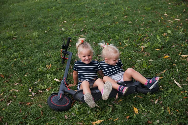 Twin blonde little girls sit on fallen electric scooter in green park