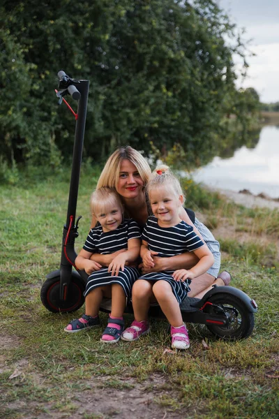 Twin blonde little girls sitting on electric scooter in park with their mother