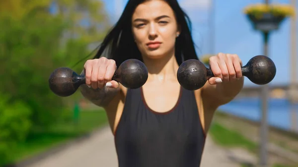 Fit brunette woman, blurry face, holds two old dumbbells during sunny day and looks at you