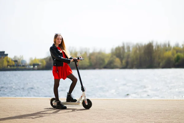 Ung kvinna i läder jacka och klänning håller elektrisk skoter — Stockfoto