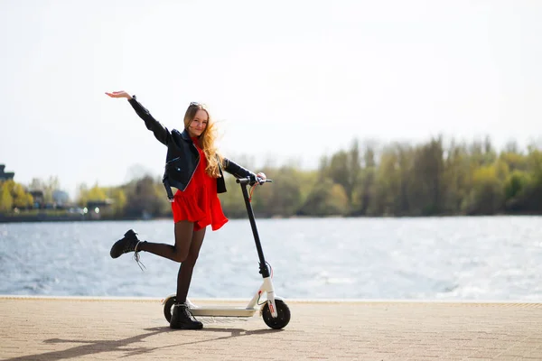 Gelukkig en positief blond meisje verhoogt hand en been in de buurt van elektrische scooter — Stockfoto