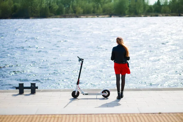 Vrouw in lederen jas staat in de buurt van elektrische scooter op zonnige promenade achterzijde uitzicht — Stockfoto