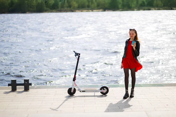Jonge blonde vrouw staat in de buurt van witte elektrische scooter op de promenade met koffie — Stockfoto