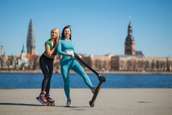 Glada blonda och brunett systrar har kul med rullskridskor och elektrisk skoter på stadens strandpromenad — Stockfoto