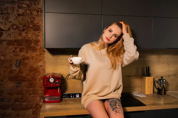 Mulher se senta na mesa, segura xícara de café e olha para a câmera de manhã — Fotografia de Stock