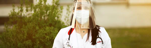 Banner van jonge vrouwelijke arts in plastic schild en chirurgische masker zitten met laptop en kijken naar je — Stockfoto