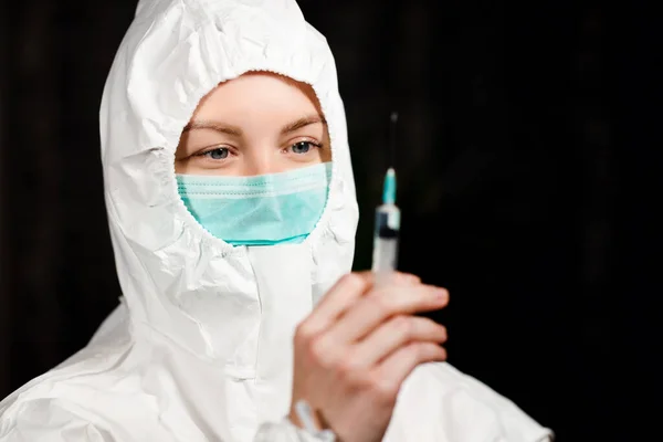 Лікар в захисному медичному костюмі та хірургічній масці дивиться на шприц — стокове фото