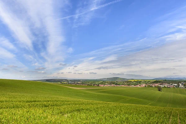 Golvende groene veld met bewolkte blauwe hemel en klein dorp — Stockfoto